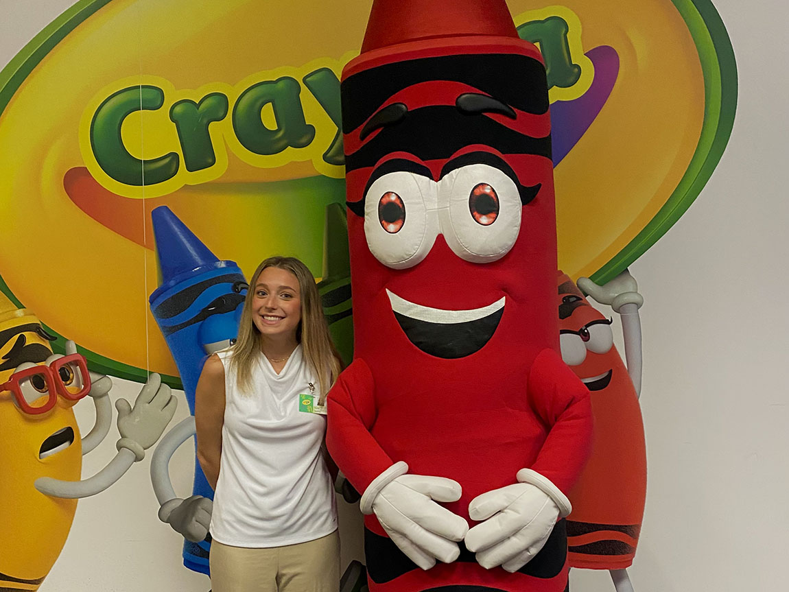 A college student in a white shirt stands next to a person dressed as a red crayon in front of the Crayola logo